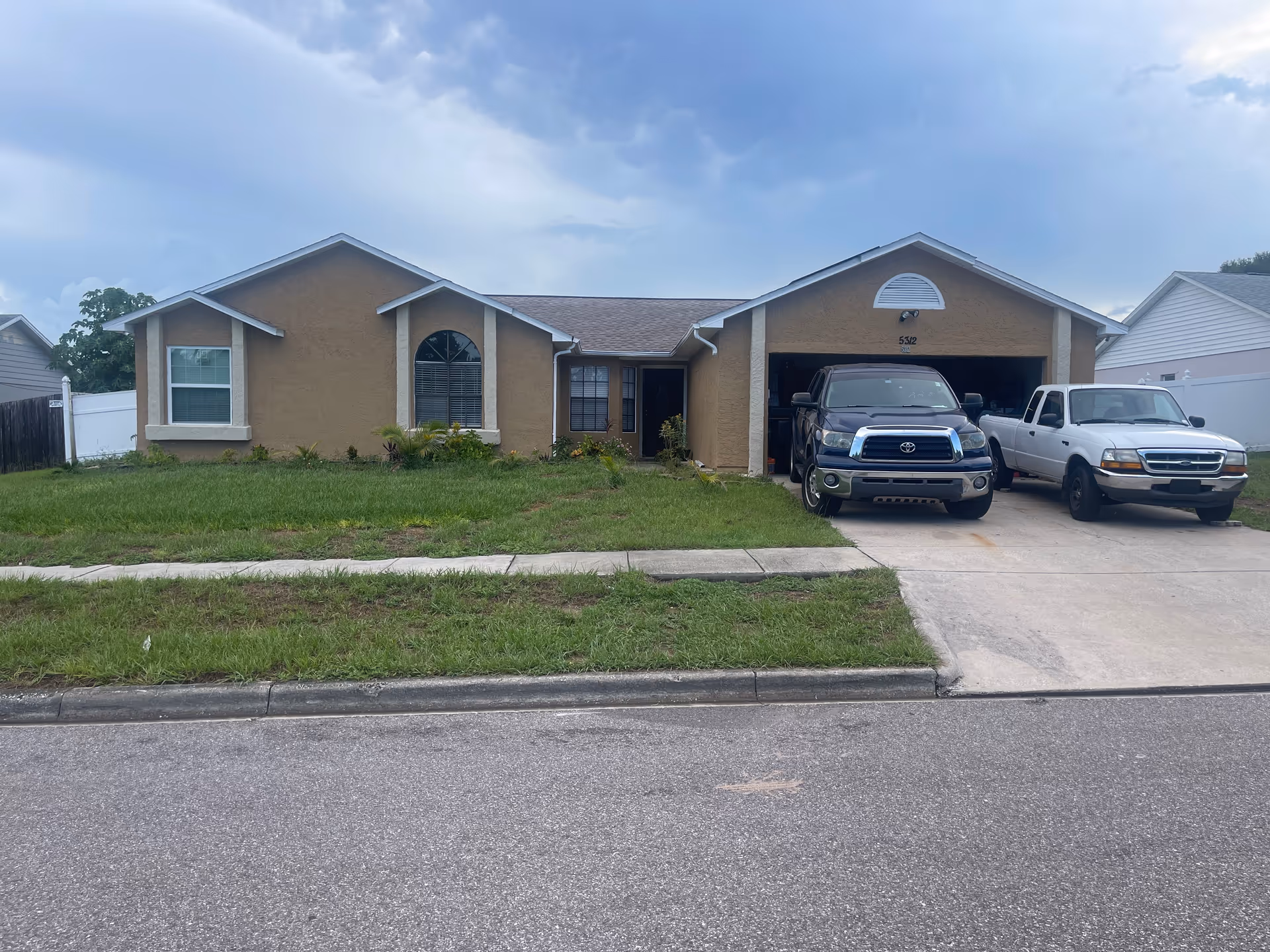 Single-story residential house with a brown exterior, two large front windows, a garage with two parked trucks, and a grassy front yard under a cloudy sky.