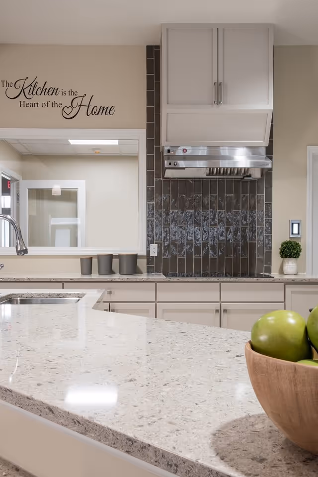 Modern kitchen interior with a light-colored granite countertop, a stainless steel sink and faucet, white cabinets, a dark tiled backsplash, and a stainless steel range hood. On the wall, there is a decorative sign that reads 'The Kitchen is the Heart of the Home'. A wooden bowl with green apples is placed on the countertop.