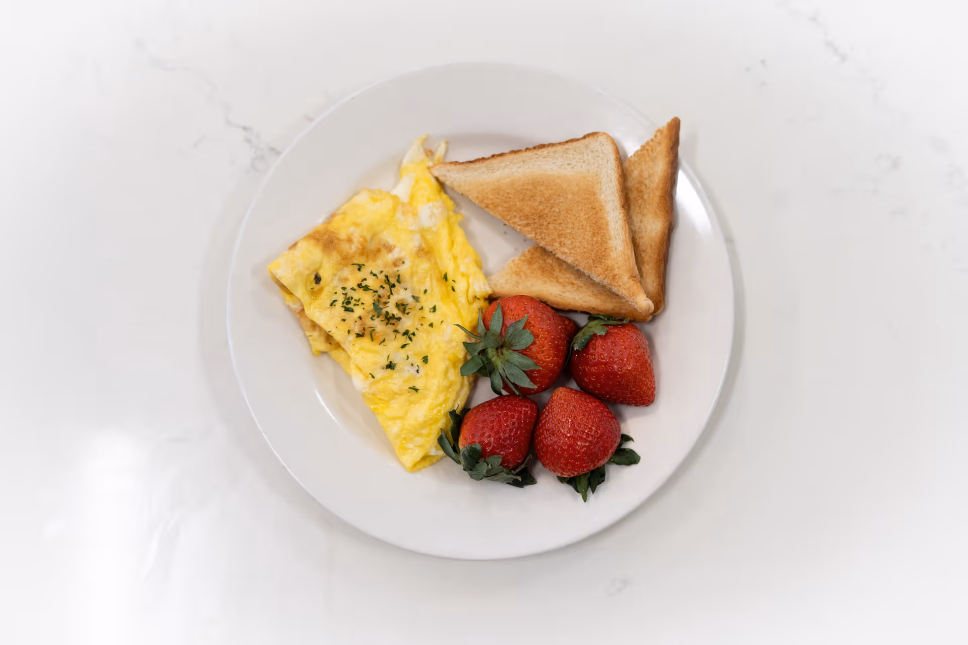A white plate with a folded yellow omelette garnished with herbs, two triangular pieces of toasted bread, and four fresh strawberries on a white marble surface.