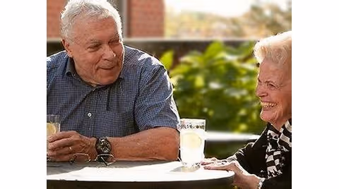An elderly man and woman sitting at an outdoor table, smiling and enjoying a conversation with glasses of lemonade in front of them. The background shows greenery and a brick structure.