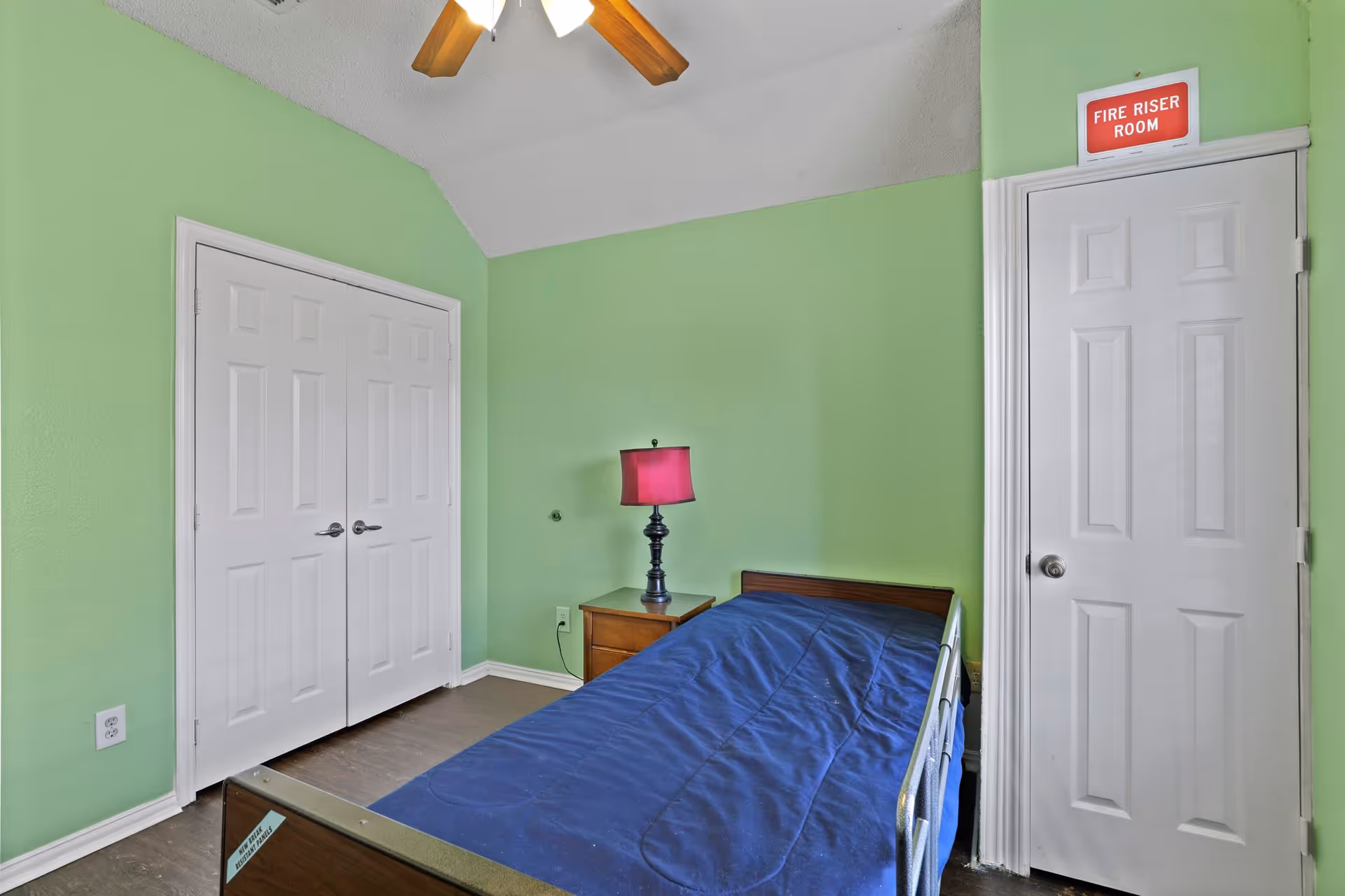 A small bedroom with light green walls, a ceiling fan with wooden blades, a single bed with a blue blanket, a wooden nightstand with a lamp that has a pink lampshade, a double white closet door, and a white door labeled 'Fire Riser Room'.