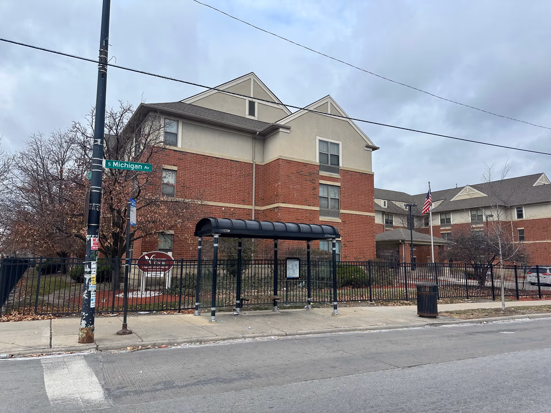 Front exterior of a three-story brick senior living building with a fenced entrance, nearby bus shelter, and an American flag.