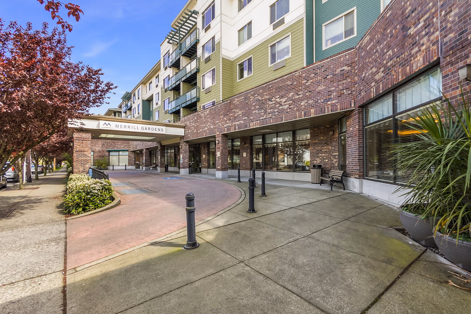 Exterior view of Merrill Gardens at Renton Centre showing the building entrance with a covered drop-off area, brick and green siding facade, sidewalk, trees with red leaves, and potted plants.