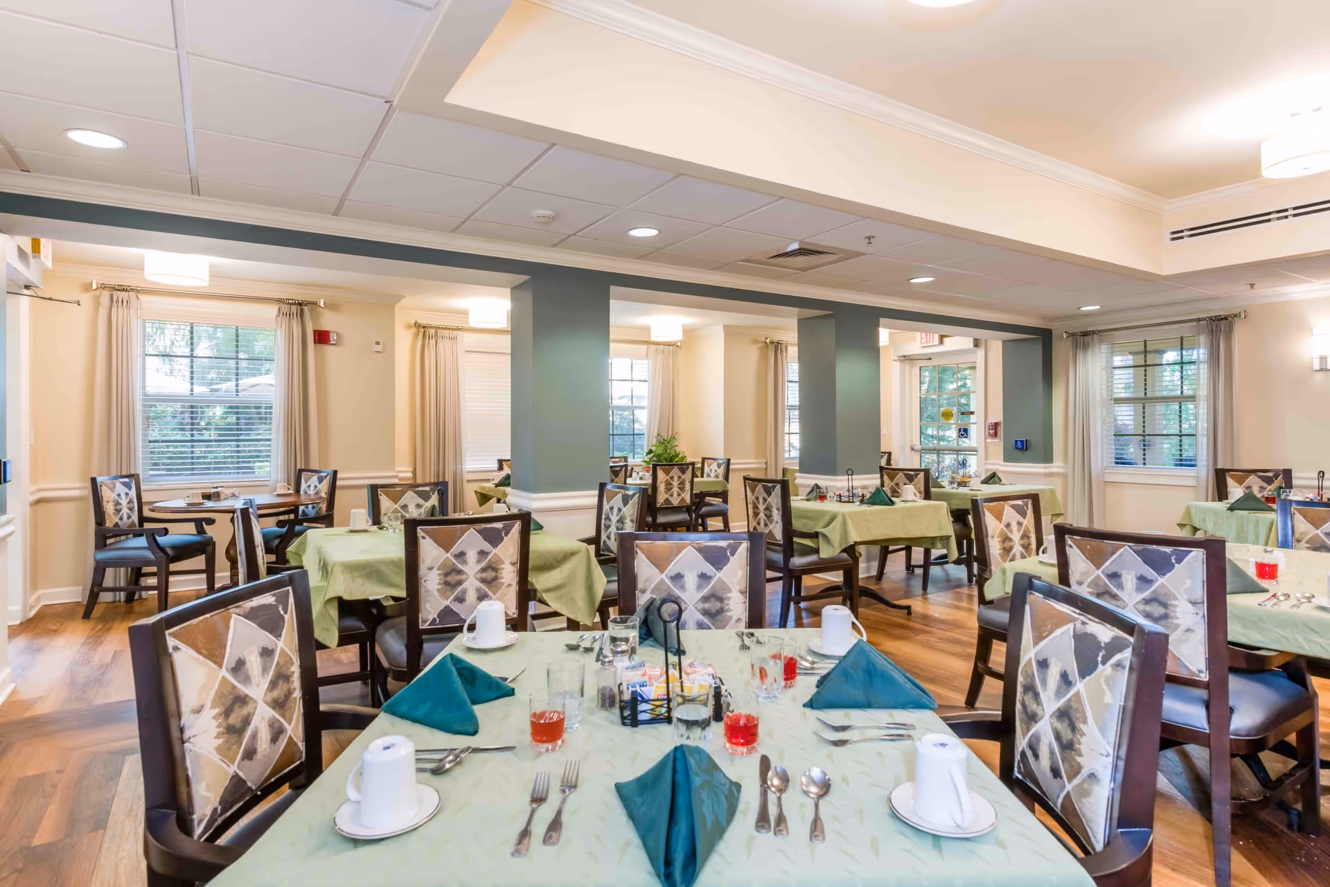 Spacious dining room with multiple tables covered in green tablecloths and set with napkins and place settings.