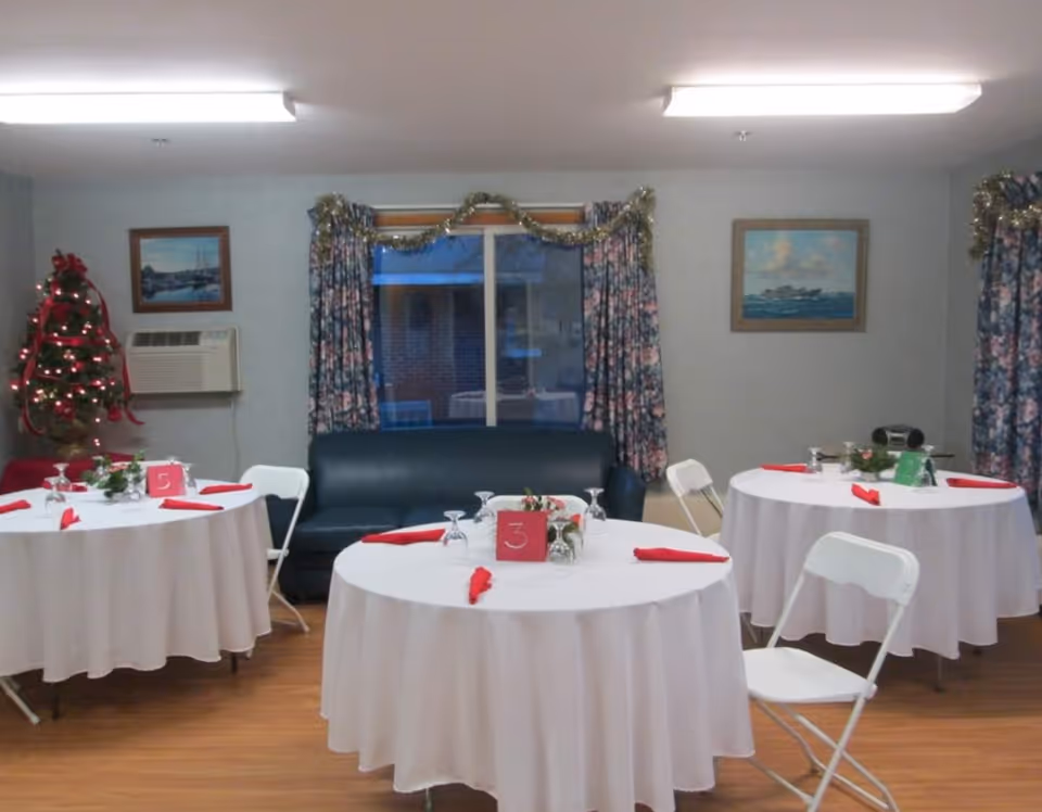 A dining room with three round tables covered with white tablecloths and set with red napkins and glassware. Each table has a small centerpiece and a number card. There are white folding chairs around the tables. A small decorated Christmas tree with red ribbons and lights is in the corner near a window with floral curtains and garland. Two framed paintings hang on the walls, and a dark blue couch is positioned against the wall under the window.