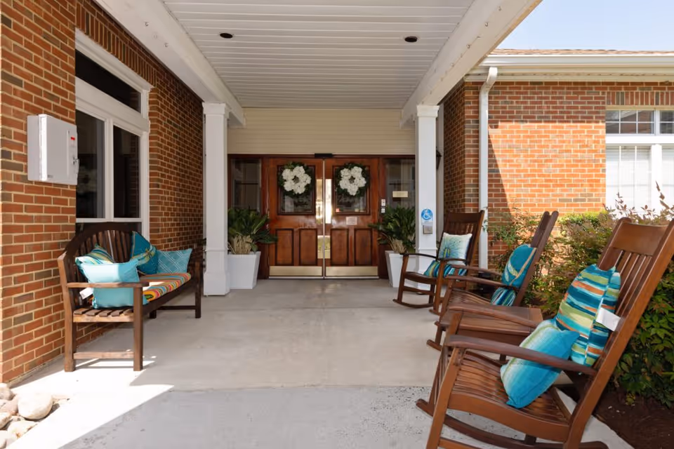 Covered entrance area of a building with double wooden doors decorated with white floral wreaths. There are wooden rocking chairs and a bench with colorful cushions arranged on either side of the entrance. The building exterior is made of red brick with white trim.