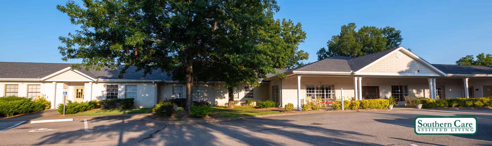 Exterior view of Southern Care Assisted Living facility showing a single-story building with a covered entrance, large windows, and surrounding greenery including bushes and a large tree in front. The parking area is visible with marked spaces and a clear blue sky above.