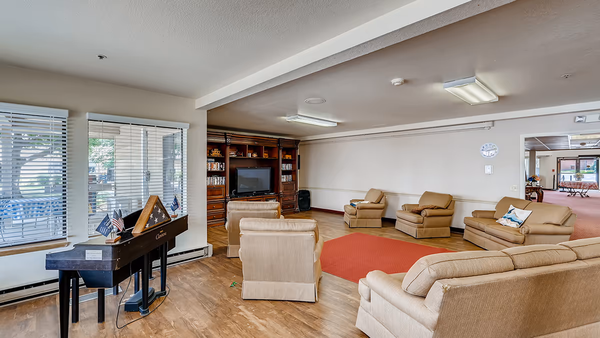 A spacious living room area with beige upholstered sofas and armchairs arranged around a red rug. There is a large wooden entertainment center with a TV and shelves filled with DVDs and decorative items. To the left, there is a piano with small American flags and a folded flag display case on top. Large windows with blinds allow natural light to enter the room.