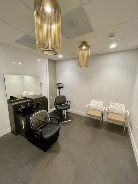 Interior view of a small salon or grooming area with two black salon chairs, a large mirror, two modern pendant lights hanging from the ceiling, and two white chairs against the wall.