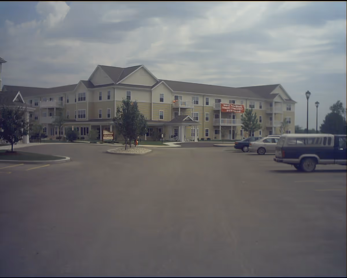 Three-story senior apartment building with balconies, a front entrance and a parking lot with a few cars under a cloudy sky.