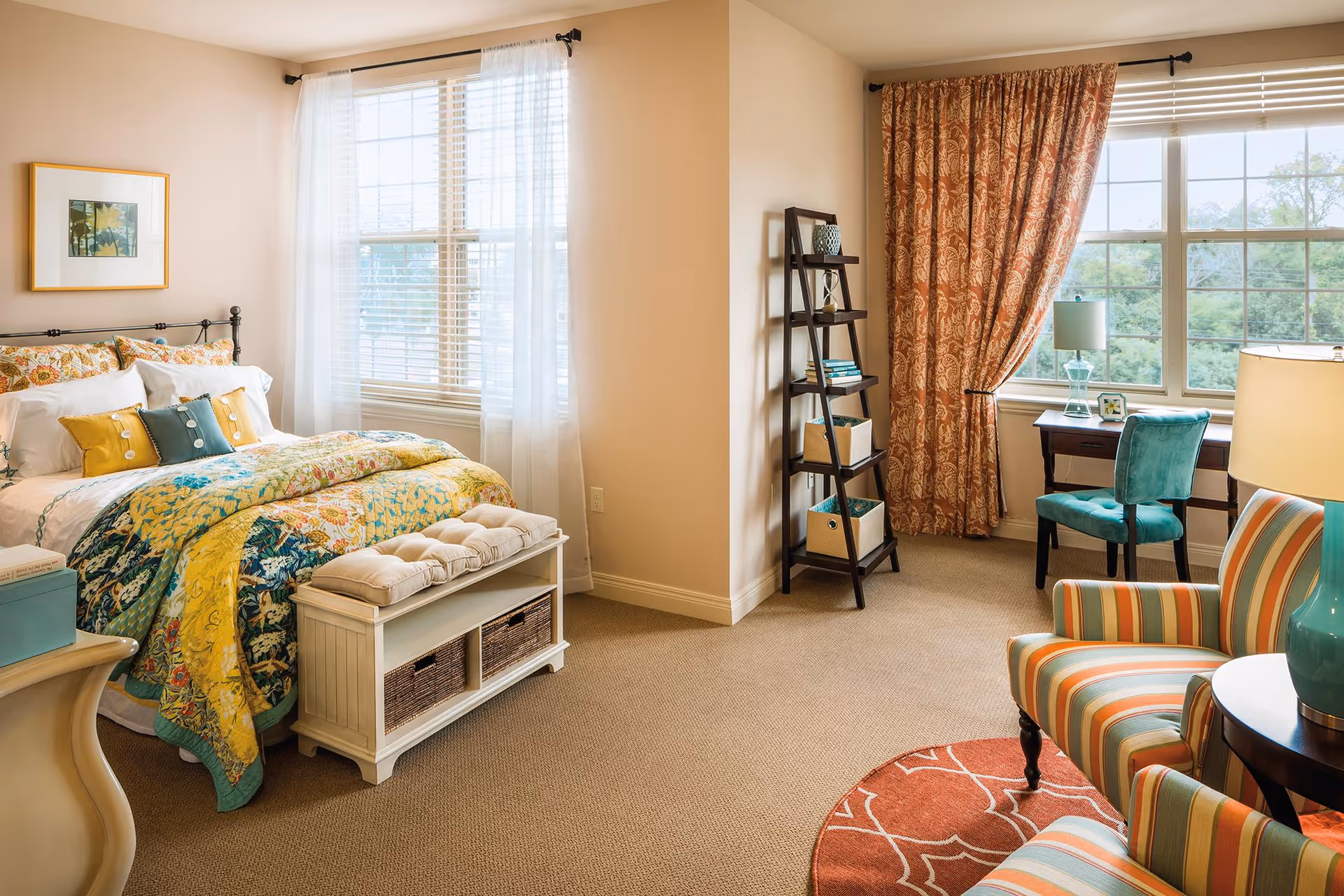 A cozy bedroom with a bed featuring colorful floral bedding and multiple pillows. The room has two large windows with sheer white and patterned orange curtains. There is a small white bench with cushions at the foot of the bed, a dark wooden ladder shelf with books and storage baskets, a desk with a turquoise chair and lamp, and two striped armchairs with a round side table and lamp. The room is carpeted and well-lit with natural light.