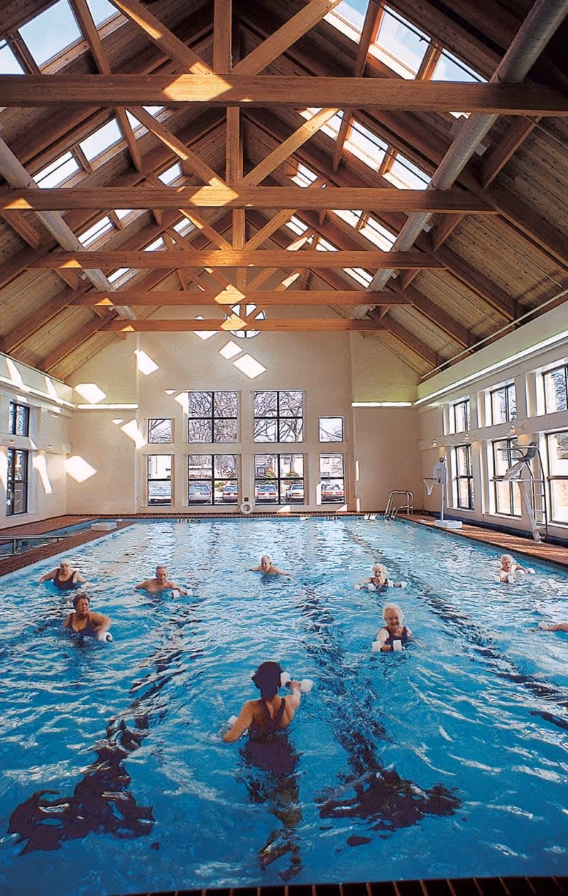 Indoor swimming pool with a wooden vaulted ceiling and large windows letting in natural light. Several elderly people are participating in a water exercise class, using foam dumbbells in the pool.