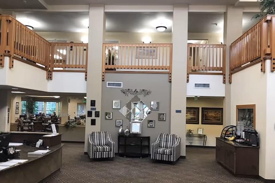 Interior view of a senior living facility lobby or common area with a high ceiling and wooden railings on the upper level. Two striped armchairs are placed against a wall decorated with framed pictures and a diamond-shaped mirror. To the left is a reception desk, and to the right is a small refreshment station with a water dispenser and snacks. In the background, there is a dining area with tables and chairs.