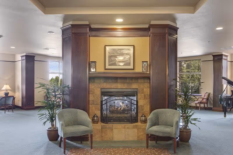 A cozy senior living facility common area with a central fireplace surrounded by dark wood paneling and tile. Two green upholstered chairs are placed in front of the fireplace, flanked by potted plants. The room has large windows letting in natural light, additional seating areas, and a grand piano visible on the right side.