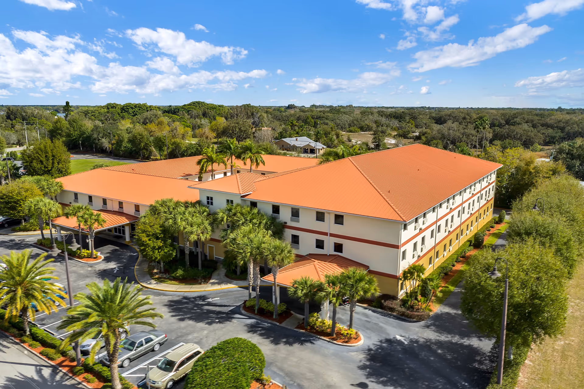 Aerial view of Balmoral Assisted Living facility showing a large, three-story building with a red roof surrounded by palm trees and greenery under a blue sky with scattered clouds. There is a parking lot with several cars in front of the building.