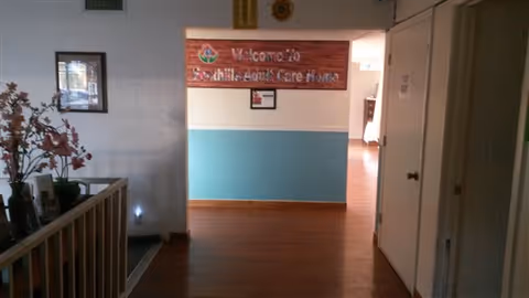 Interior hallway of Foothills Adult Care Home with wooden flooring and a blue and white painted wall. A wooden sign on the far wall reads 'Welcome to Foothills Adult Care Home'. There is a framed picture on the left wall and a small table with flowers and other items near a railing.