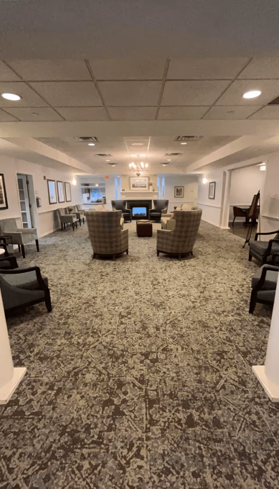 A spacious, well-lit common area with patterned carpet flooring, several upholstered armchairs arranged around a small ottoman, and a fireplace with a painting above it. The room has a chandelier, recessed ceiling lights, framed artwork on the walls, and additional seating along the sides.