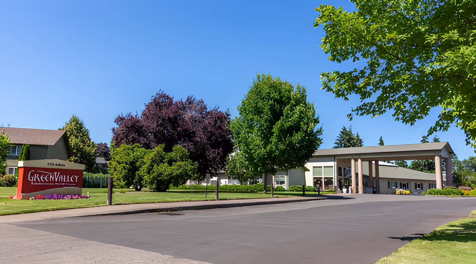 Exterior view of Green Valley Rehabilitation Health Center on a clear sunny day, showing the building entrance with a covered driveway, surrounded by green trees and a well-maintained lawn with a red sign displaying the facility's name.