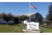 Single-story residential care home building with a well-maintained lawn, several trees, and an American flag on a flagpole. A white sign in front reads 'Holiday House'.