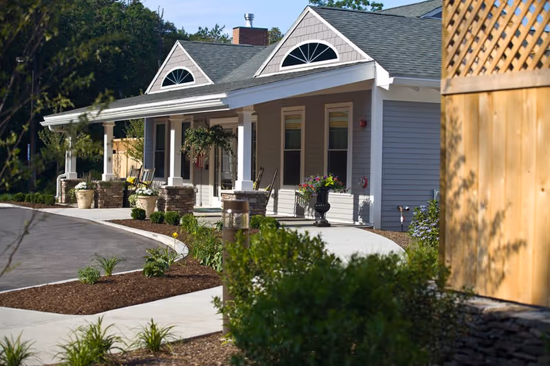 Exterior view of a single-story building with a covered porch featuring white columns and stone bases. The building has gray siding, two decorative half-circle windows above the porch, and a landscaped area with plants and mulch along a curved sidewalk. There are chairs and potted plants on the porch, and a wooden fence is visible on the right side.