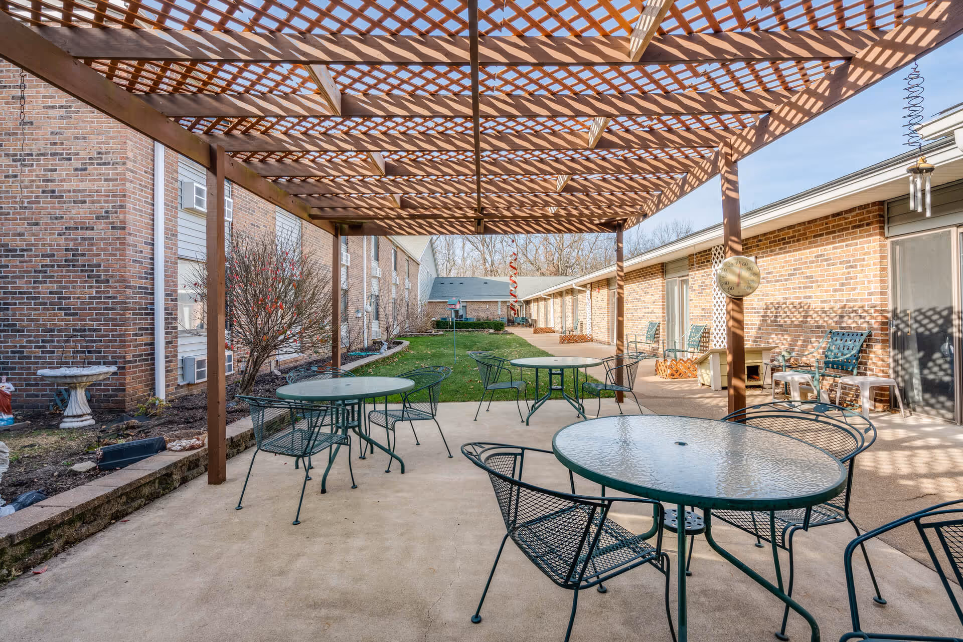 Covered outdoor courtyard with metal patio tables and chairs under a wooden pergola beside a brick senior living building and grassy walkway.