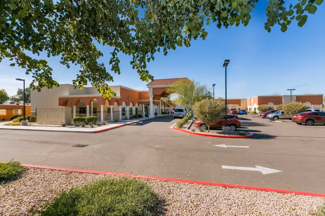 Front entrance of a single-story senior living facility with a covered drive-through, landscaped parking lot, and several parked cars under a clear blue sky.