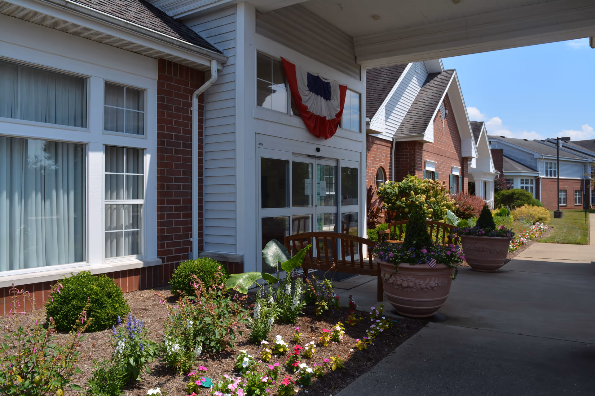 Exterior view of The Glenwood Community building entrance with a garden featuring colorful flowers and shrubs, two large flower pots with plants, wooden benches, and a patriotic red, white, and blue banner above the doorway under a clear blue sky.