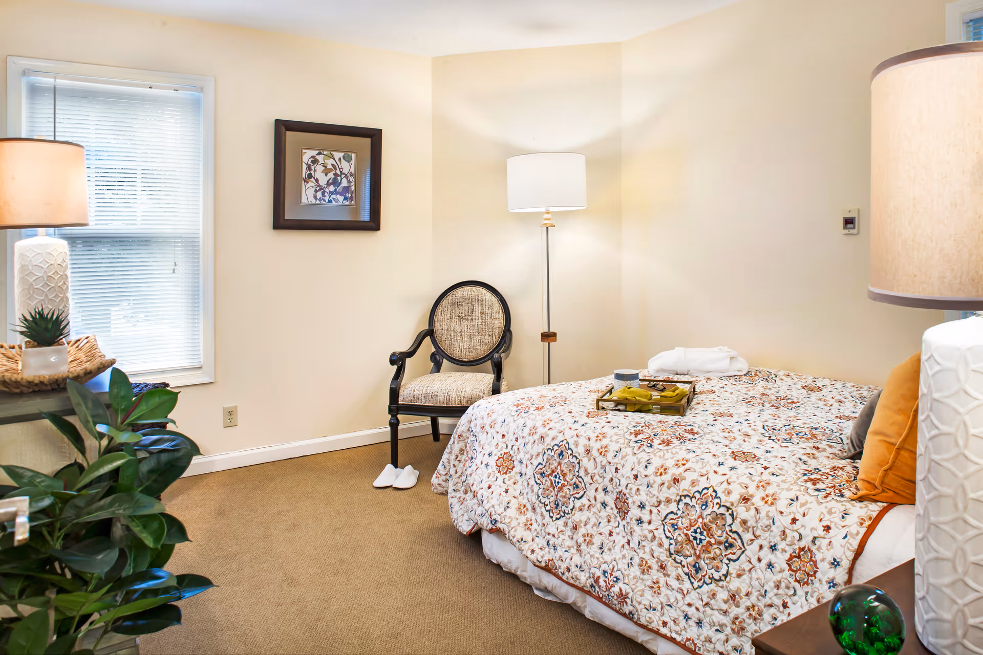 A cozy bedroom with a bed covered in a floral patterned quilt, a cushioned armchair, a floor lamp, two table lamps, a window with blinds, and a green plant in the corner.