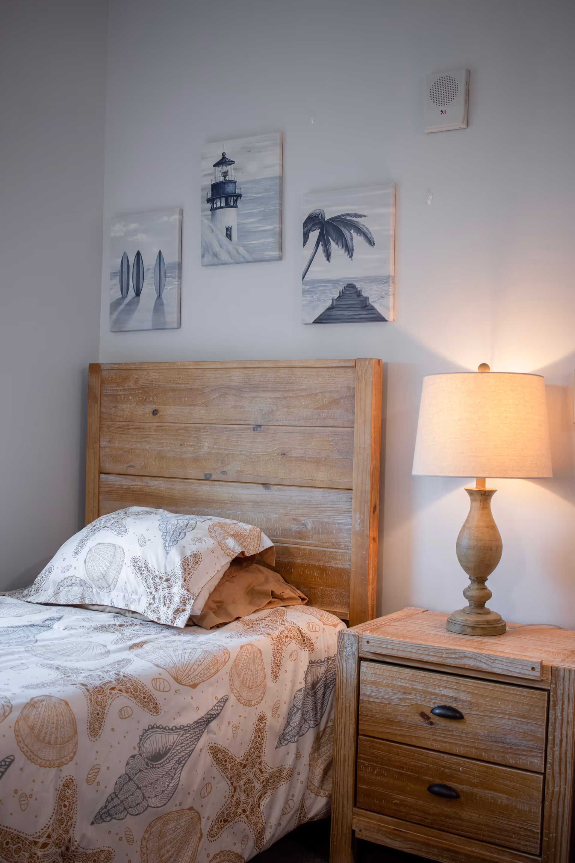A cozy bedroom with a wooden headboard and matching wooden nightstand. The bed is made with seashell and starfish patterned bedding. On the nightstand is a lamp with a beige shade, casting a warm light. Above the bed are three framed pictures depicting a lighthouse, surfboards, and a palm tree with a pier, all in muted tones.