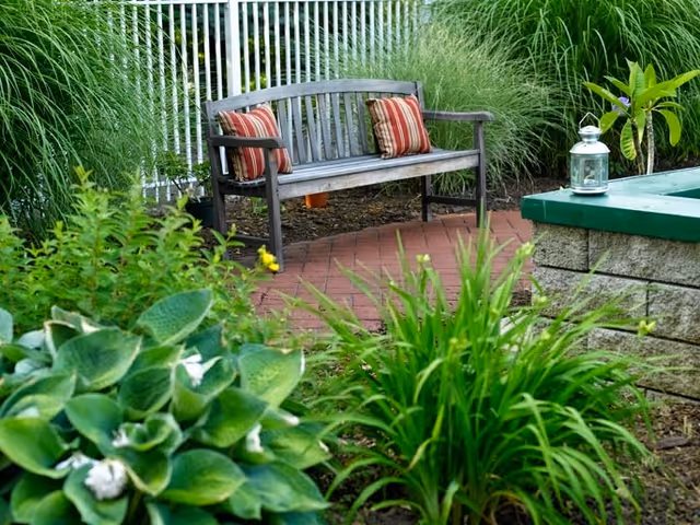 A wooden bench with two striped cushions placed on a brick patio surrounded by green plants and tall grasses, with a white fence in the background and a small lantern on a stone ledge nearby.