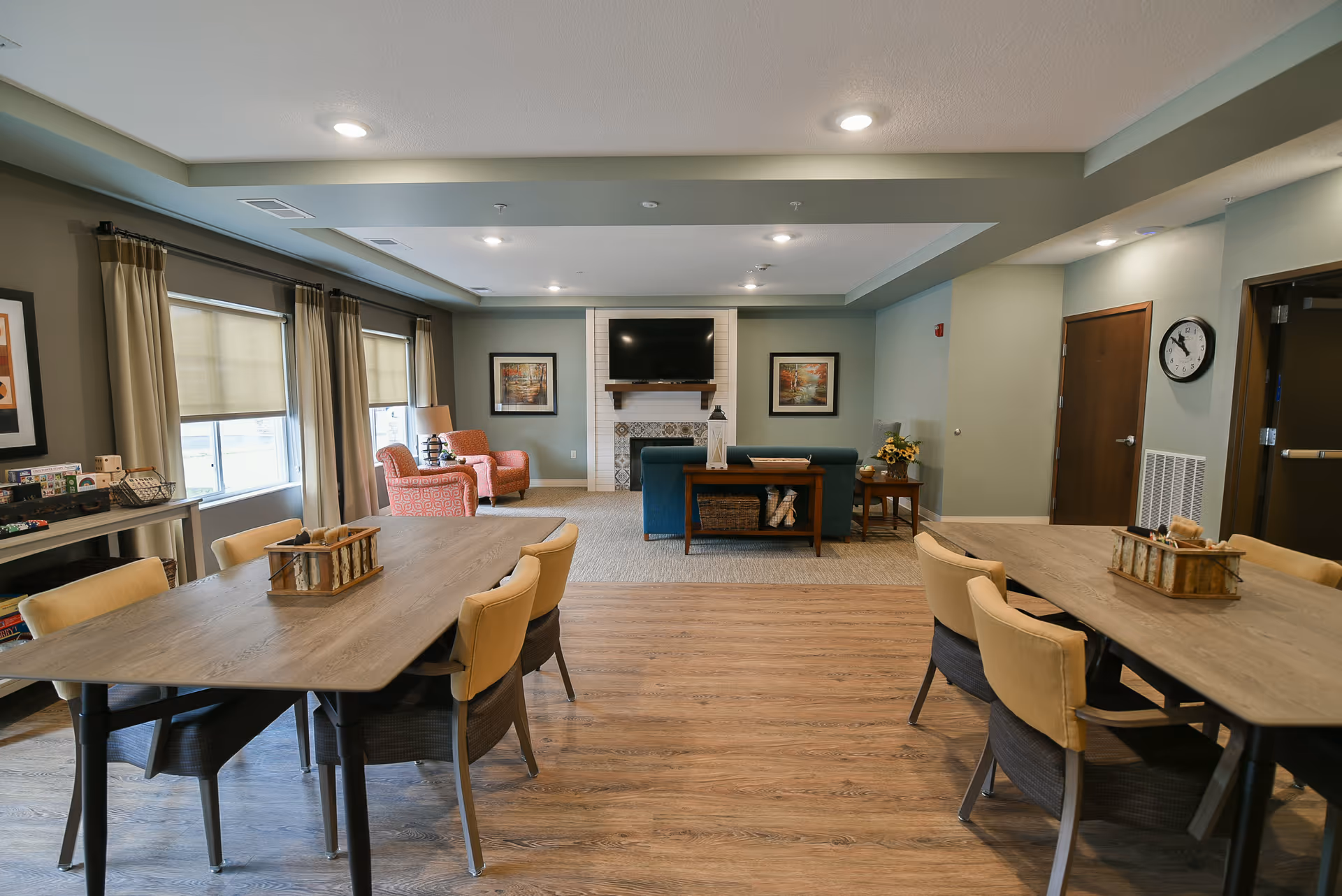 A spacious living room area in a senior living facility with two wooden tables surrounded by beige chairs in the foreground. In the background, there is a seating area with a blue sofa, two orange armchairs, a fireplace with a mounted TV above it, and framed artwork on the walls. The room has large windows with beige curtains and wood flooring.