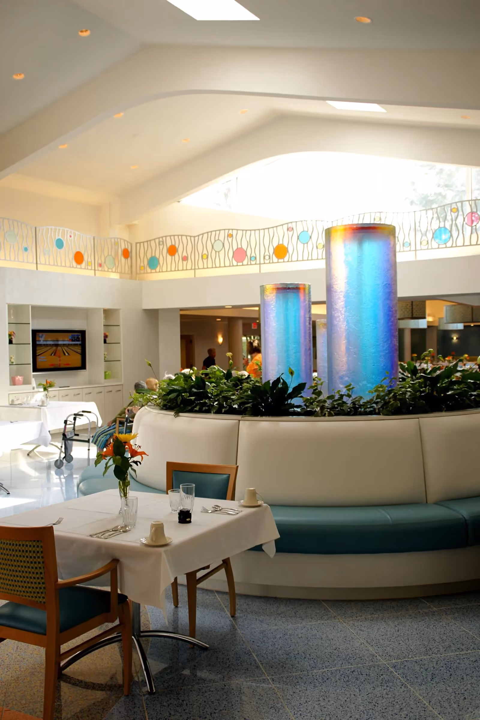 A bright and spacious dining area in a senior living facility with tables set with white tablecloths, chairs, and a centerpiece of flowers. In the center is a circular seating area with plants and two tall, colorful illuminated columns. The room has a high ceiling with skylights and a decorative railing with colorful circular accents on the upper level.