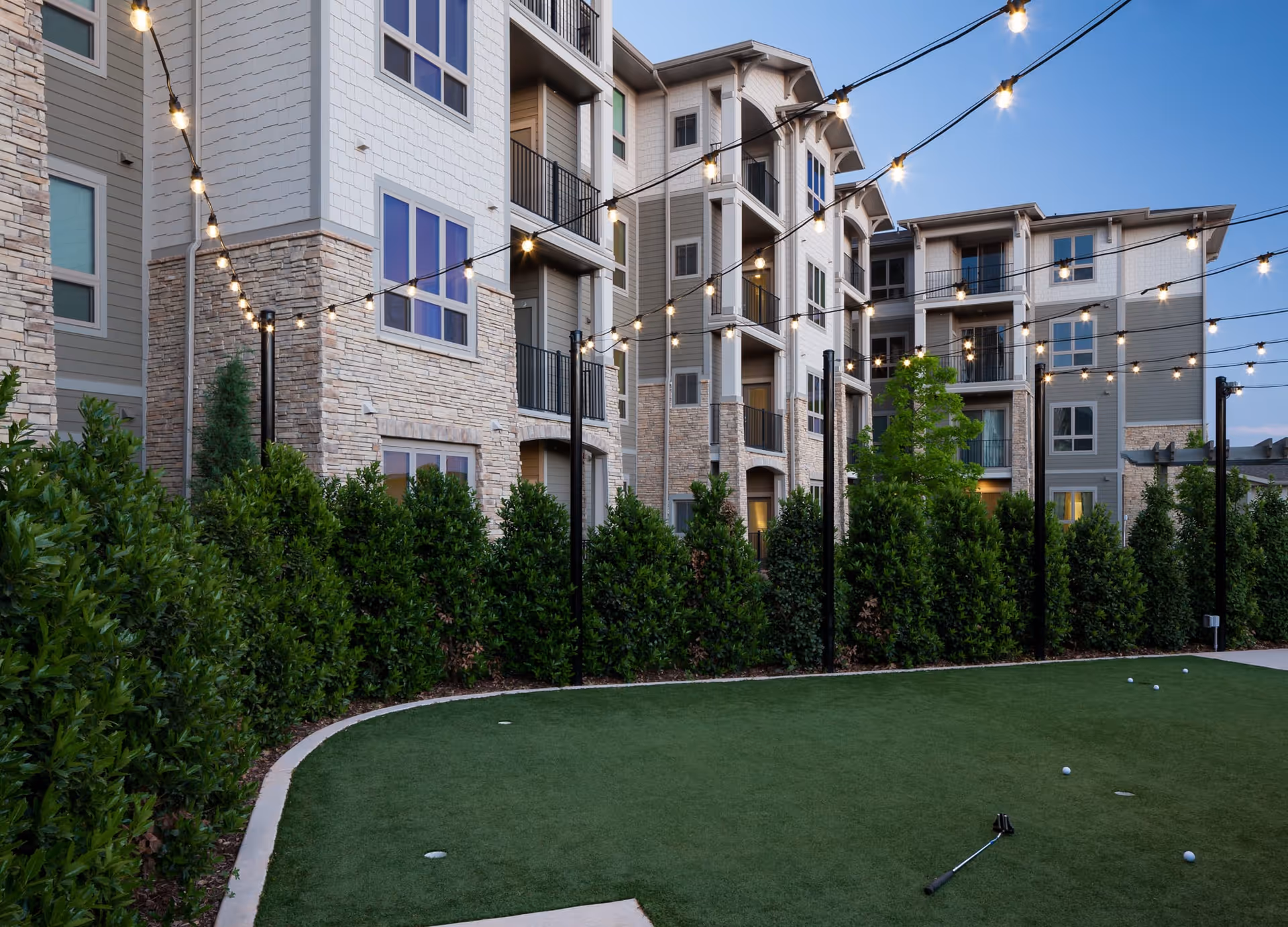Outdoor putting green area with golf balls and a putter, surrounded by tall green bushes and a multi-story residential building with balconies and string lights overhead during dusk.