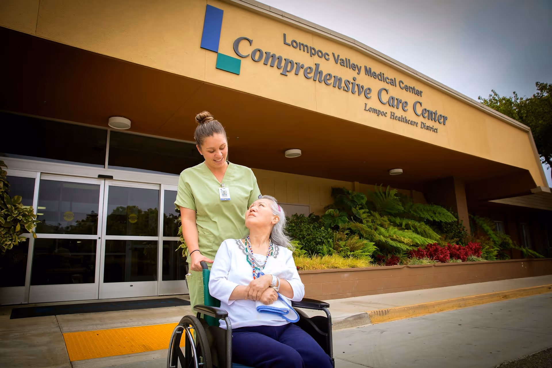 A healthcare worker in green scrubs is standing behind an elderly woman in a wheelchair outside the entrance of Lompoc Valley Medical Center Comprehensive Care Center. The elderly woman is looking up at the healthcare worker, and there are plants and shrubs along the building's exterior.