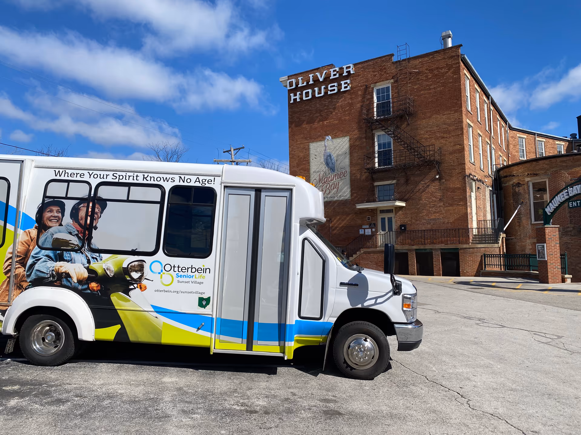 A white shuttle bus with colorful blue, green, and yellow stripes parked in front of a large brick building labeled 'Oliver House'. The bus has an image of two smiling elderly people riding a scooter and the text 'Where Your Spirit Knows No Age!' along with the Otterbein SeniorLife Sunset Village logo and website. The sky is blue with some clouds.