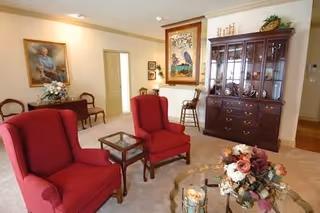 A cozy living room with two red upholstered armchairs facing a glass coffee table adorned with a floral centerpiece and candles. In the background, there is a wooden china cabinet with glass doors displaying decorative items, a framed painting on the wall, and a small table with a flower arrangement. The room has beige walls and carpeted flooring.
