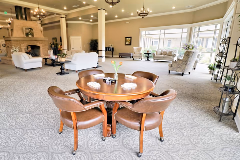 Spacious senior living common lounge with a round wooden table and four leather chairs in the foreground, sofas and a fireplace in the background and large windows letting in natural light.