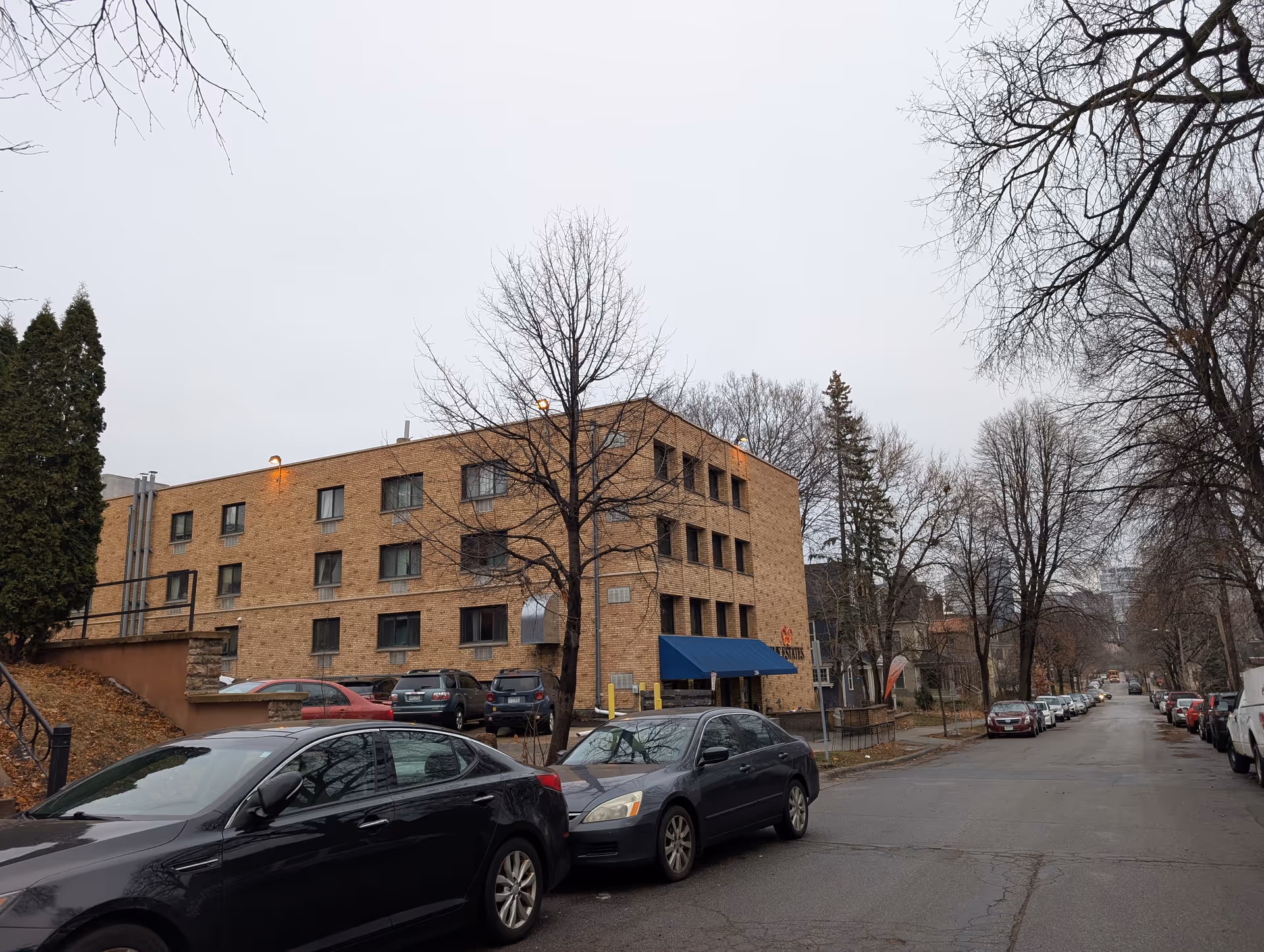 Three-story brick building with a blue awning on a residential street lined with parked cars and leafless trees.