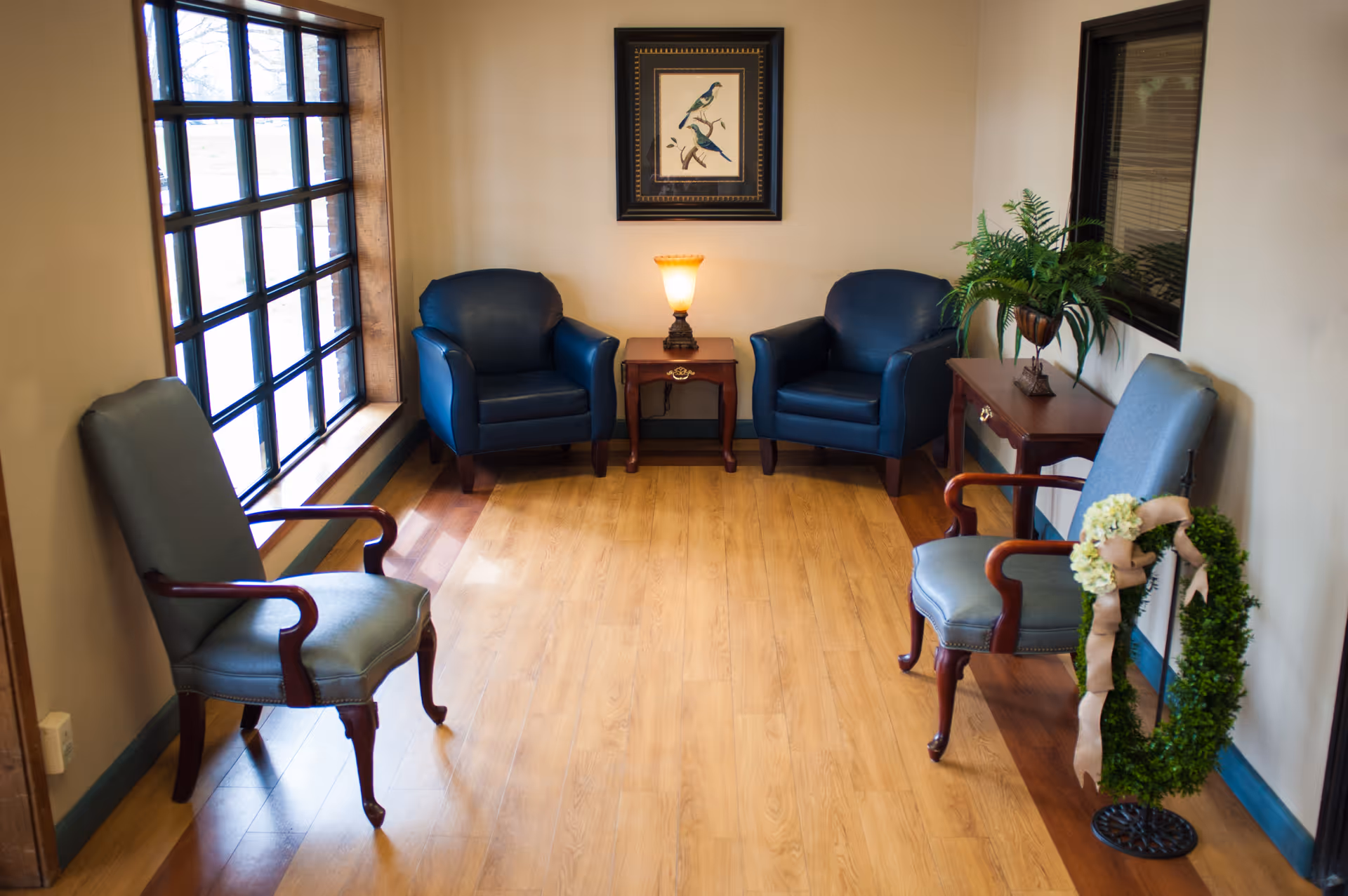 A small sitting area with four chairs arranged around a wooden floor. Two blue leather armchairs are placed against the far wall with a small wooden side table and a lamp between them. Two upholstered chairs with wooden armrests are positioned on either side near windows. A framed picture of birds hangs on the wall above the lamp, and a green plant in a pot sits on a wooden console table to the right. A decorative wreath with white flowers and a ribbon is placed on a stand near the right chair.