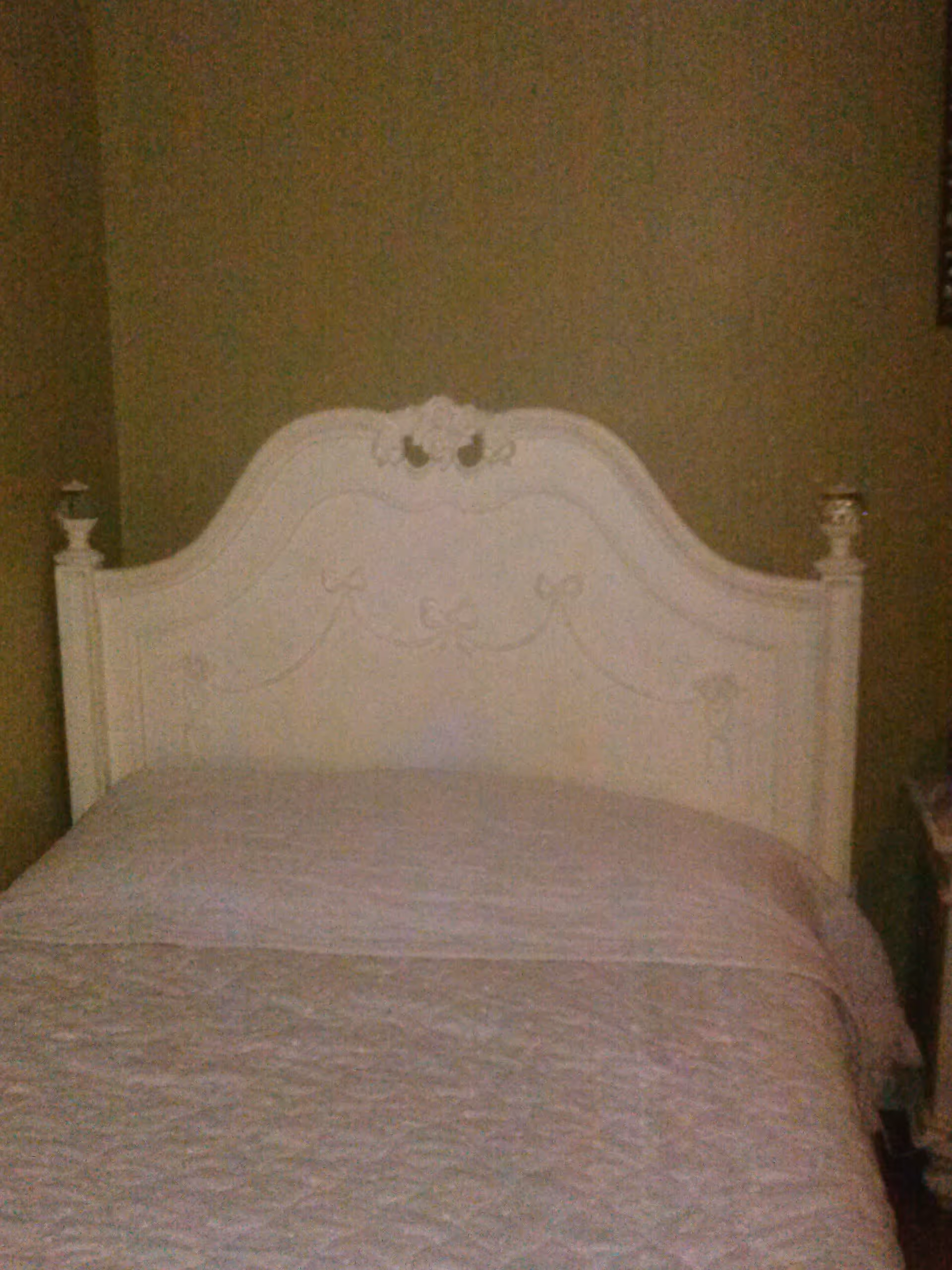 A bedroom with a white ornate headboard bed against a beige wall. The bed is made with a light-colored quilted bedspread.