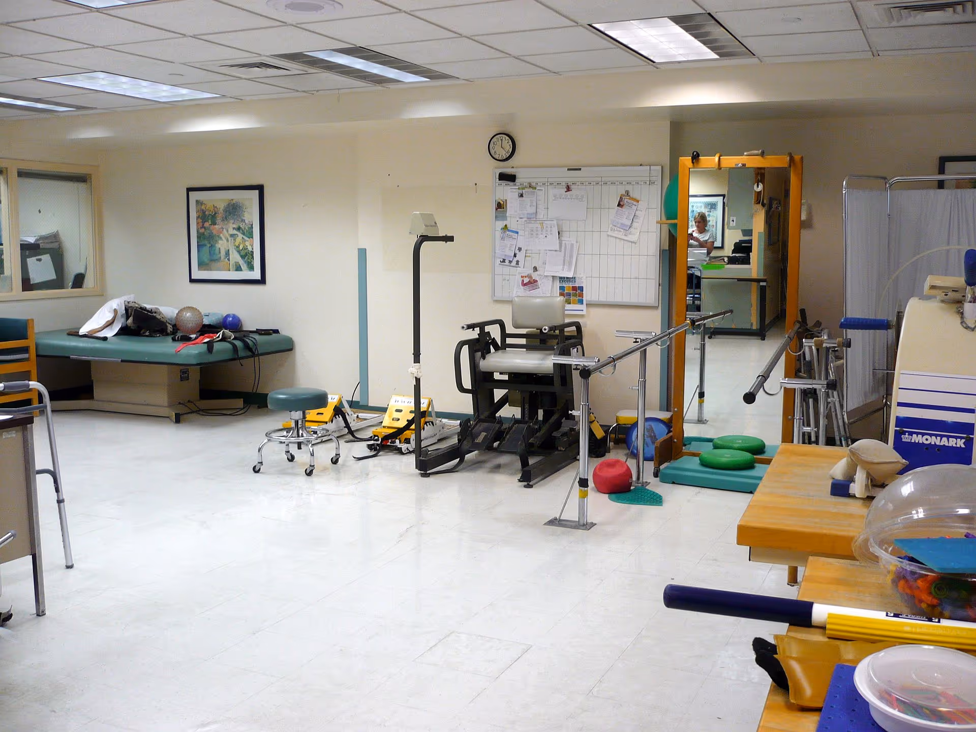 A physical therapy or rehabilitation room in a nursing home with exercise equipment including a leg press machine, parallel bars for walking practice, therapy balls, a padded treatment table, and various therapy tools. There is a clock and a whiteboard with papers on the wall, a large mirror reflecting part of the room, and a window into another room.