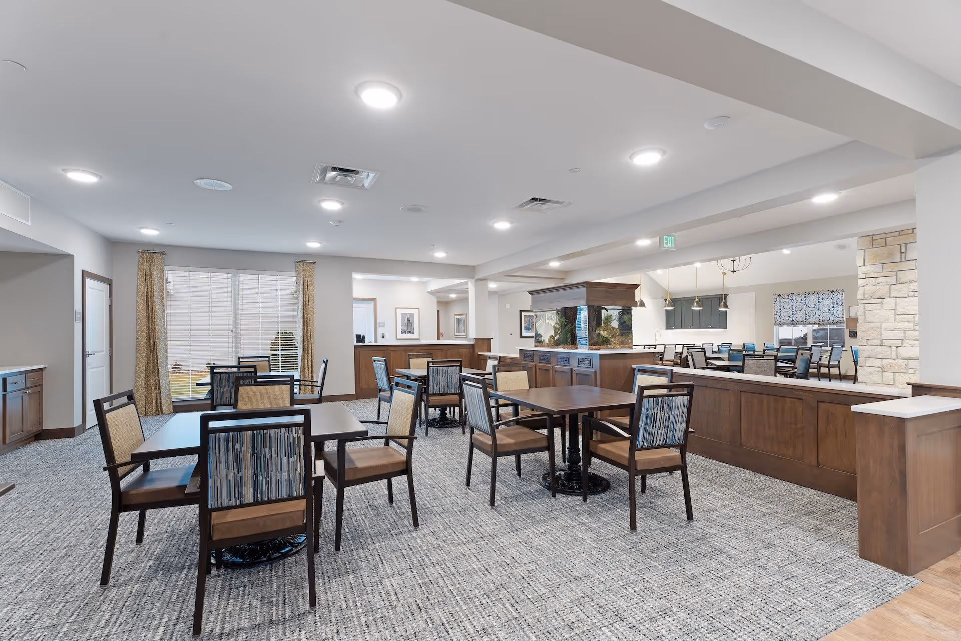 A spacious senior living dining area with multiple wooden tables and chairs arranged on a patterned carpet. The room features a large fish tank centerpiece, neutral-colored walls, and bright ceiling lights. There are large windows with curtains and an adjacent room with additional seating visible in the background.
