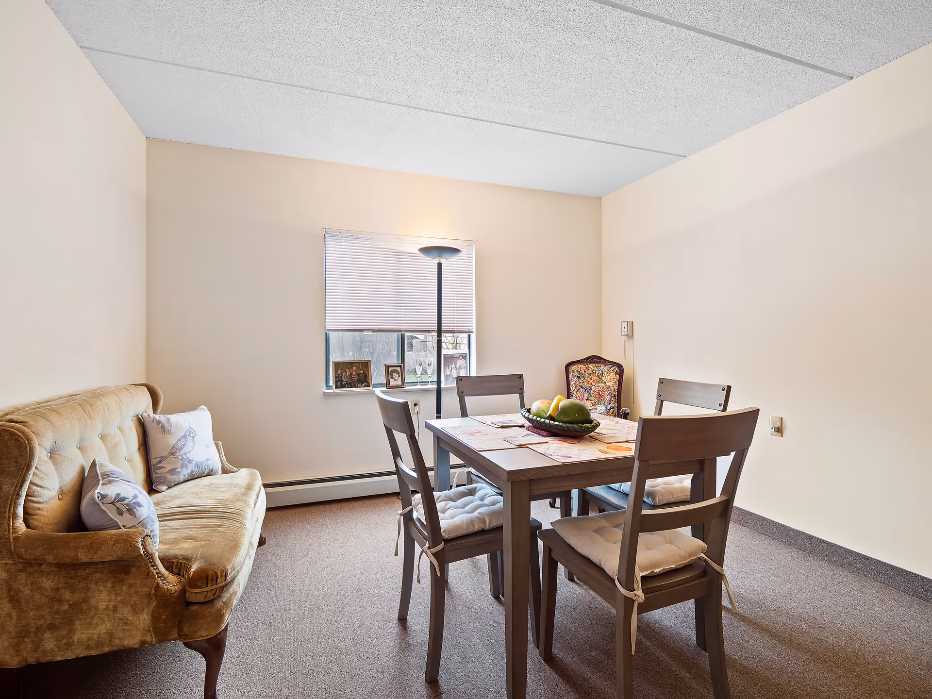 Small dining room with a wooden table and four chairs, a cushioned sofa to the left, and a window with a floor lamp.