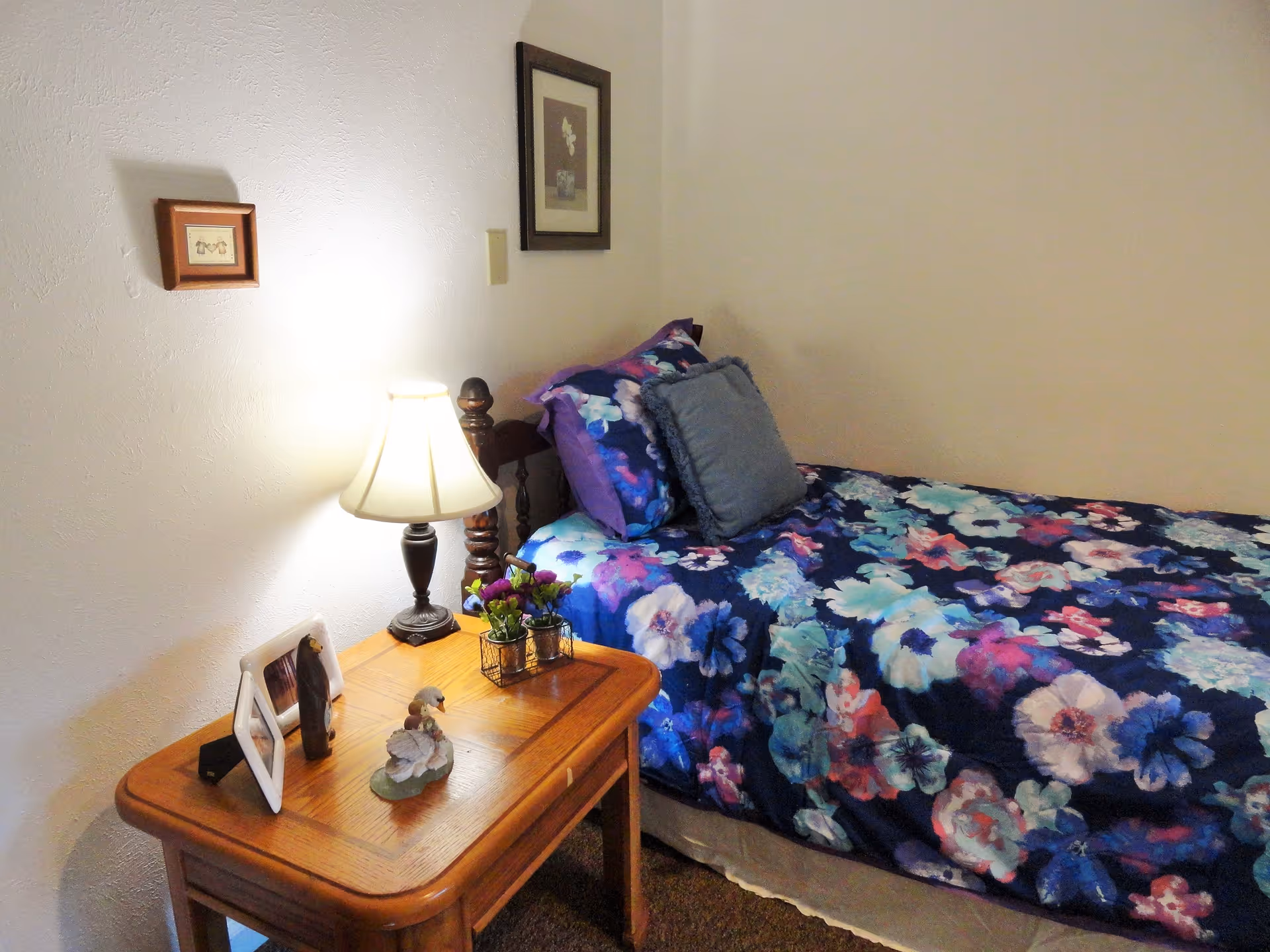 A cozy bedroom with a single bed covered in a colorful floral bedspread and matching pillows. Next to the bed is a wooden nightstand with a lit table lamp, small framed pictures, a figurine, and a small flower arrangement. Two framed pictures hang on the wall above the bed and nightstand.
