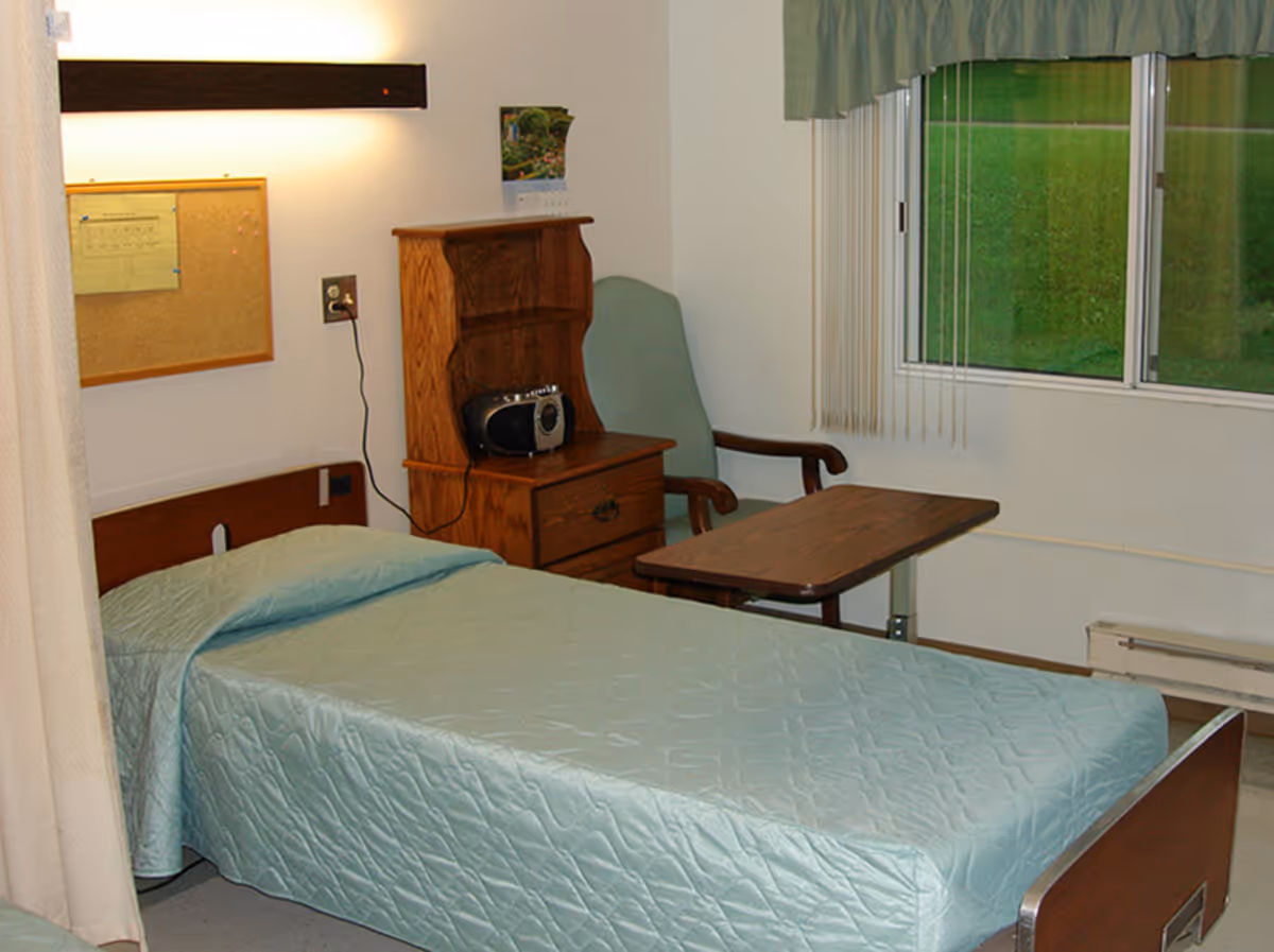 A small senior-care bedroom with a single bed covered in a light blue quilt, a wooden nightstand and chair, an overbed table, and a window with vertical blinds.