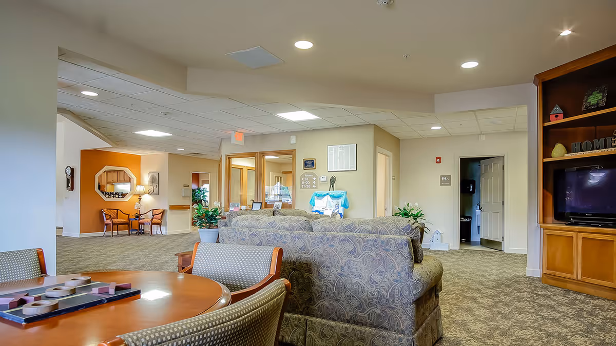 A spacious and well-lit common area in Stonegate Village Assisted Living & Memory Care featuring patterned sofas, a round wooden table with chairs, a built-in wooden entertainment center with a TV, and decorative plants. The area has beige walls, carpeted floors, and multiple doorways leading to other rooms.