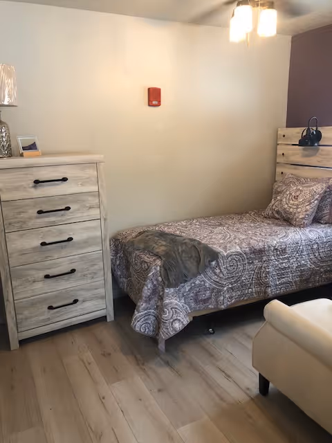 A small bedroom with a single bed covered in a patterned bedspread and matching pillow. Next to the bed is a light-colored wooden dresser with five drawers and black handles. A lamp and a small framed picture are on top of the dresser. The room has light wood flooring and a ceiling light fixture with two bulbs.