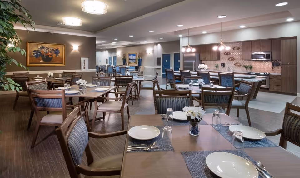 Communal dining area with multiple set tables and chairs and an open kitchen in the background.