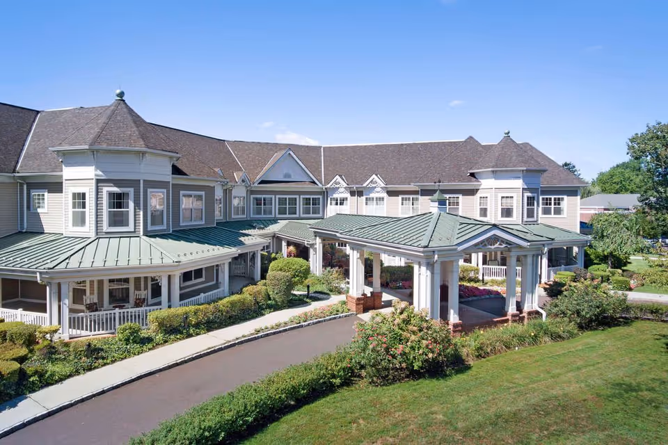 Front exterior of a two-story senior living facility with a covered porte-cochere, green metal roofs, wraparound porch, and landscaped grounds.