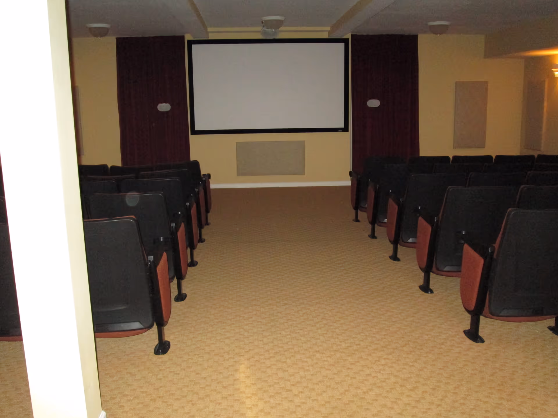 Interior view of a small theater or screening room with rows of black and red cushioned chairs facing a large blank projection screen mounted on a beige wall with dark red curtains on either side.
